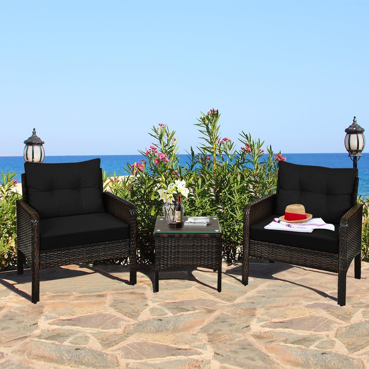 3-Piece Black Rattan Outdoor Patio Conversation Set with Cushions & Glass Table displayed on a patio overlooking the ocean, featuring two comfortable chairs and a stylish coffee table surrounded by greenery.
