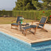Relaxing outdoor scene featuring the Patio Rattan Folding Lounge Chair with Acacia Wood Frame & Retractable Footrest by Dream Yard Oasis, accompanied by a small table and surrounded by greenery and a serene poolside.