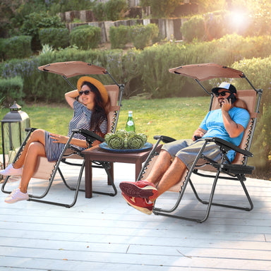 Folding Recliner Lounge Chair With Adjustable Shade Canopy & Cup Holder - Coffee, perfect for enjoying a sunny day. A man and woman relax in stylish chairs, featuring adjustable canopies, on a sunny deck.